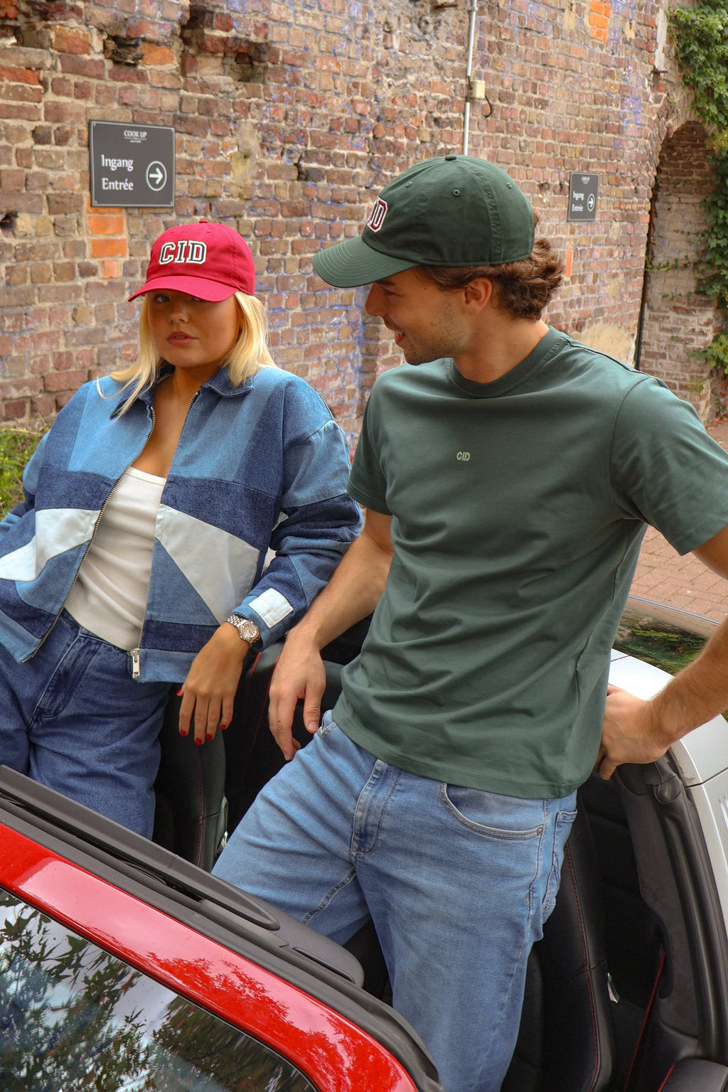 A man and a woman standing next to a red convertible car in front of a brick wall. The woman is wearing a blue patchwork denim jacket and matching jeans with a pink cap, and the man is wearing a green t-shirt, blue jeans, and a green cap.