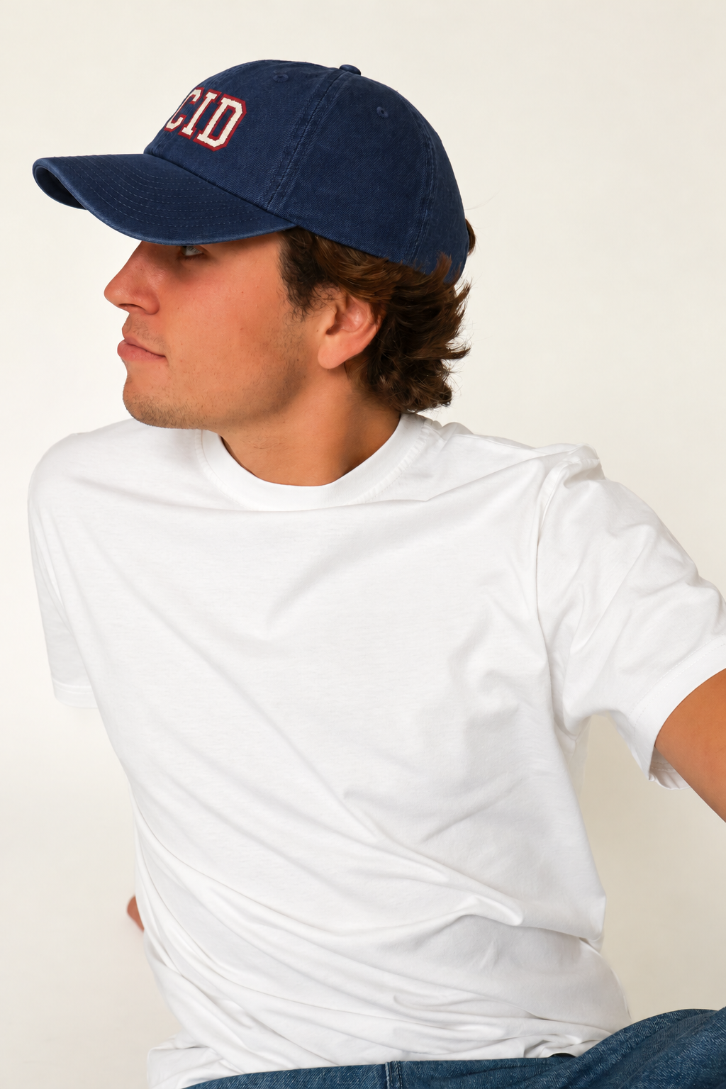 A man wearing a navy blue baseball cap with red embroidered letters and a plain white t-shirt, photographed against a light off-white background.